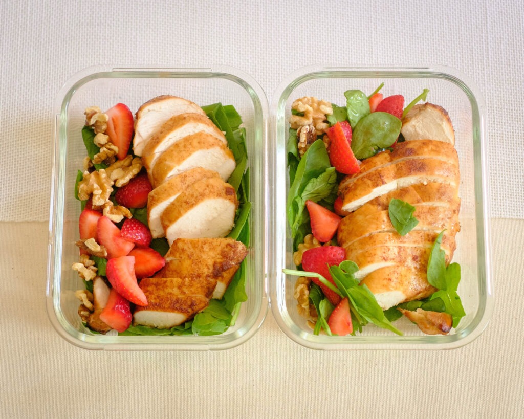 Glass meal prep containers storing baked balsamic chicken served with spinach, strawberries, and walnuts.