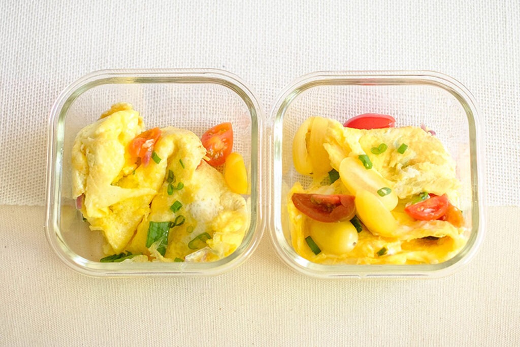 Glass meal prep containers storing a folded omelette with fresh tomatoes and green onions.