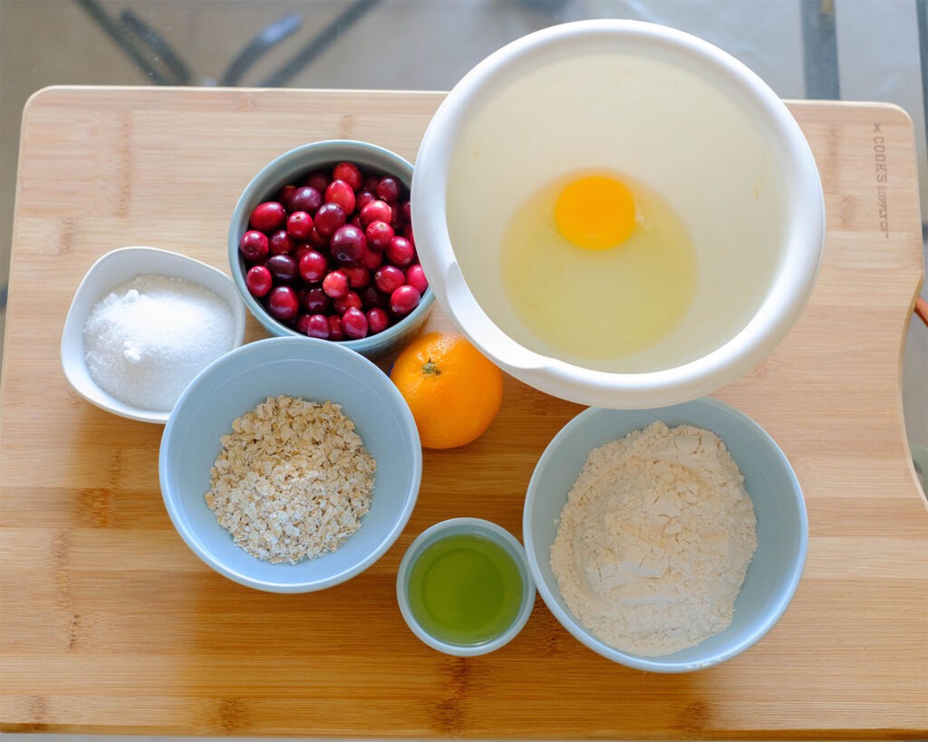 Mixing bowls that include the ingredients for cranberry muffins, such as, cranberries, egg, sugar, rolled oats, flour, orange, and olive oil