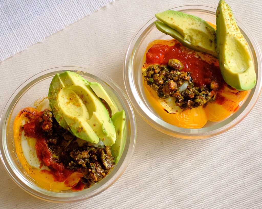 Glass meal prep containers storing a baked yellow bell pepper stuffed with eggs. It is topped off with red harissa sauce and green pesto and slice avocado.