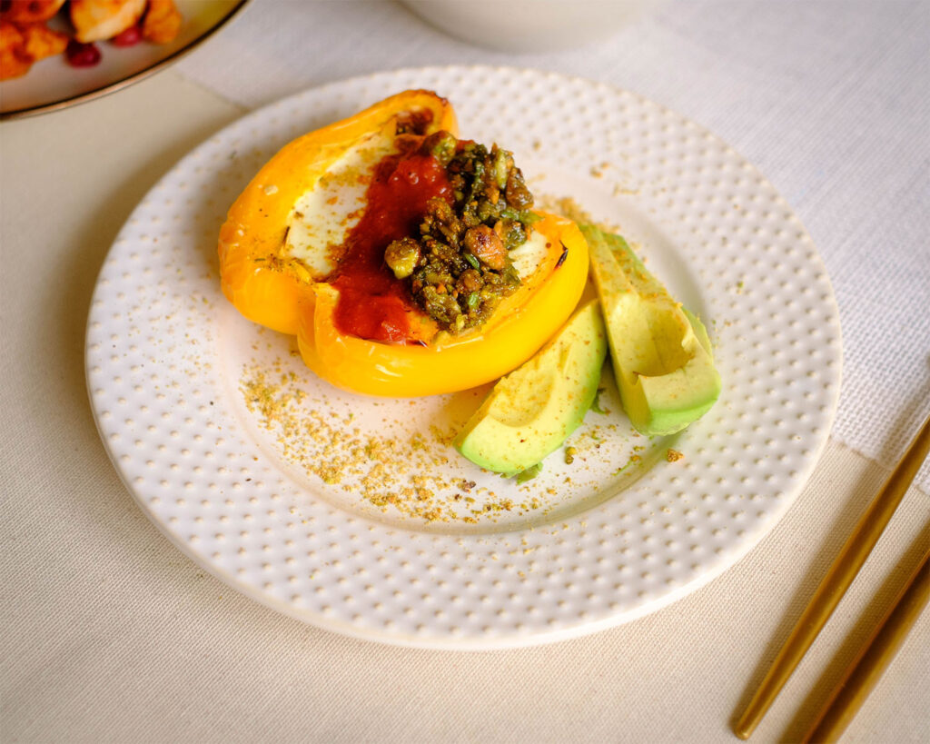 White round plate with a baked yellow bell pepper stuffed with eggs. It is topped off with red harissa sauce and green pesto and slice avocado.