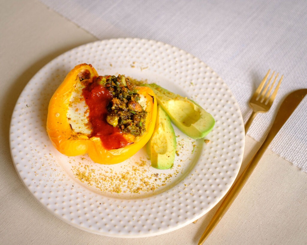White round plate with a baked yellow bell pepper stuffed with eggs. It is topped off with red harissa sauce and green pesto and slice avocado.