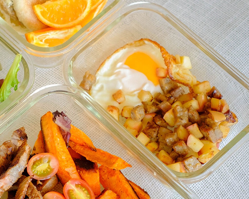 Glass meal prep containers storing a crispy fried egg served with diced apples and chicken sausage.