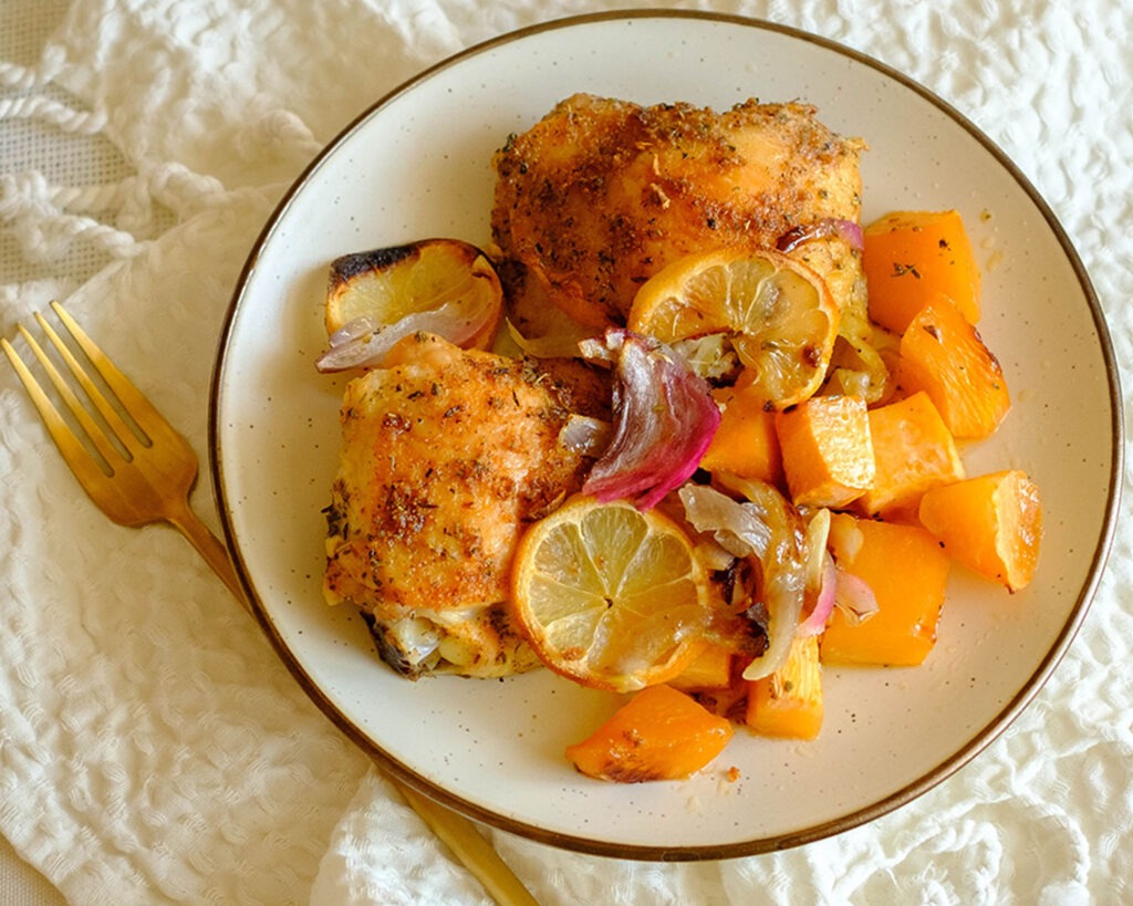 Plate with roasted chicken thighs and root vegetables, topped with lemon slices and red onions.