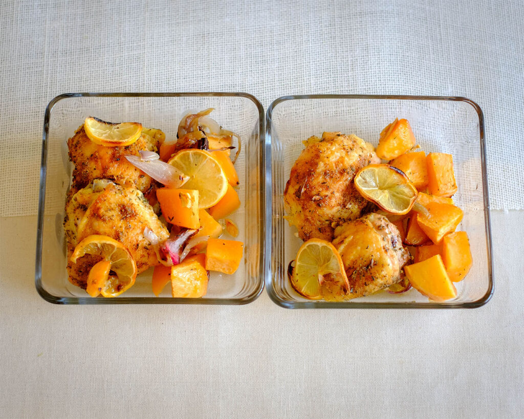 Glass meal prep containers with roasted chicken thighs and root vegetables, topped with lemon slices and red onions.