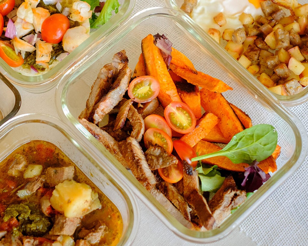 Glass meal prep container with grilled steak and sweet potato wedges on top of spring mix salad with cherry tomatoes.