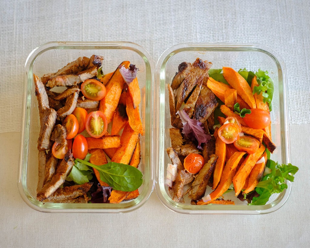 Glass meal prep containers with grilled steak and sweet potato wedges on top of spring mix salad with cherry tomatoes.