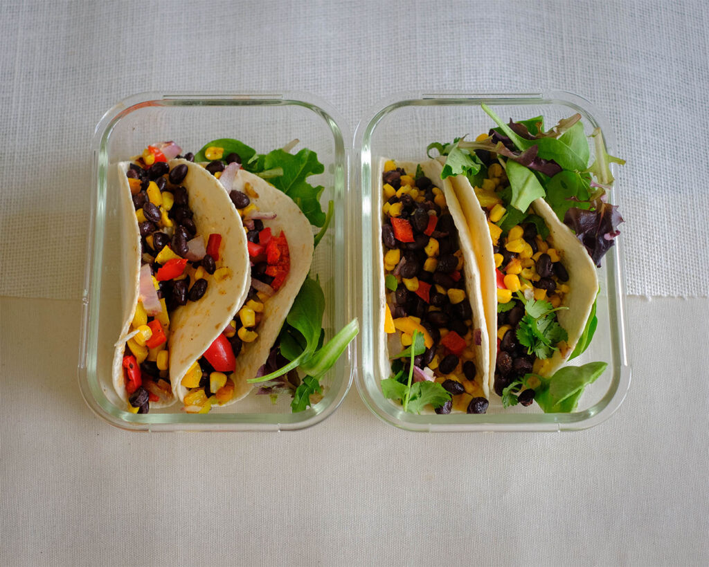 Glass meal prep containers with black bean and corn tacos with a side salad.