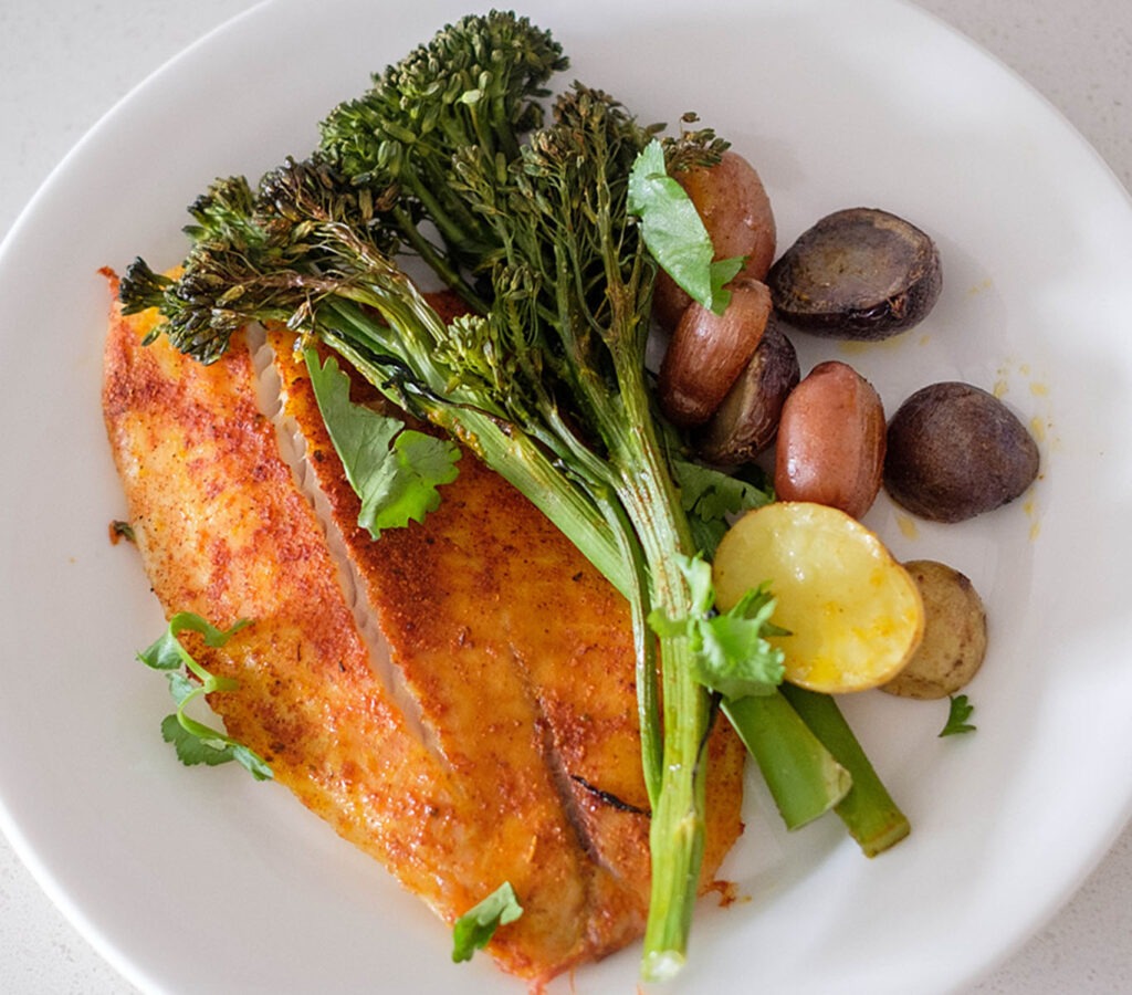 Round plate with seasoned tilapia and roasted broccolini with baby potatoes.