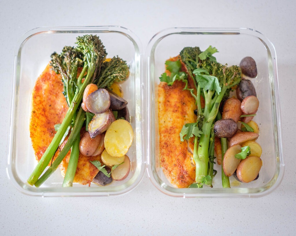 Glass meal prep containers with seasoned tilapia and roasted broccolini with baby potatoes.