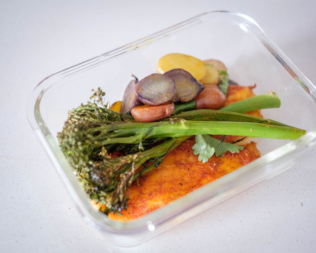 Glass meal prep container with seasoned tilapia and roasted broccolini with baby potatoes.