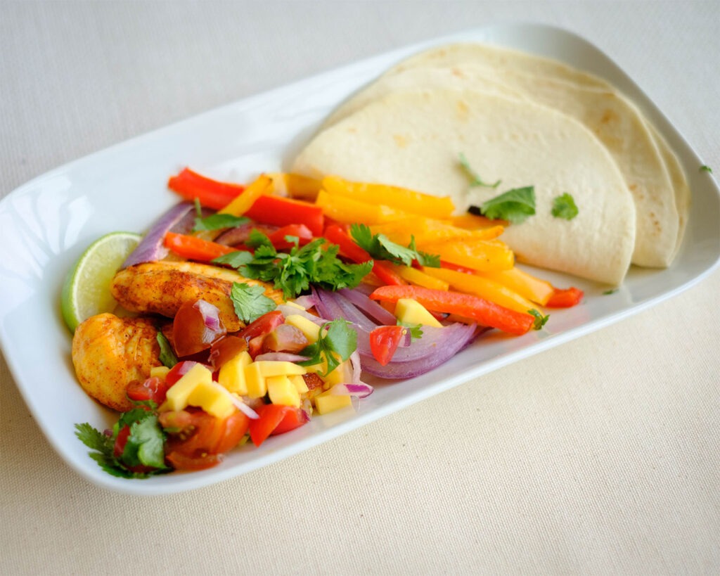 Plate with chicken fajitas, mango salsa, and colorful bell peppers and onions served with tortillas.