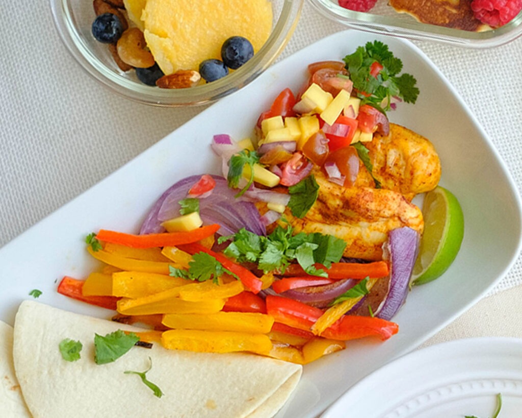 Plate with chicken fajitas, mango salsa, and colorful bell peppers and onions served with tortillas.