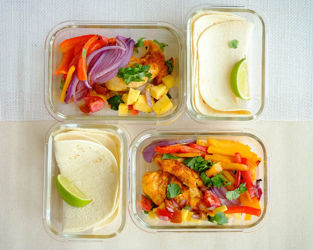 Glass meal prep containers with chicken fajitas, mango salsa, and colorful bell peppers and onions served with tortillas.