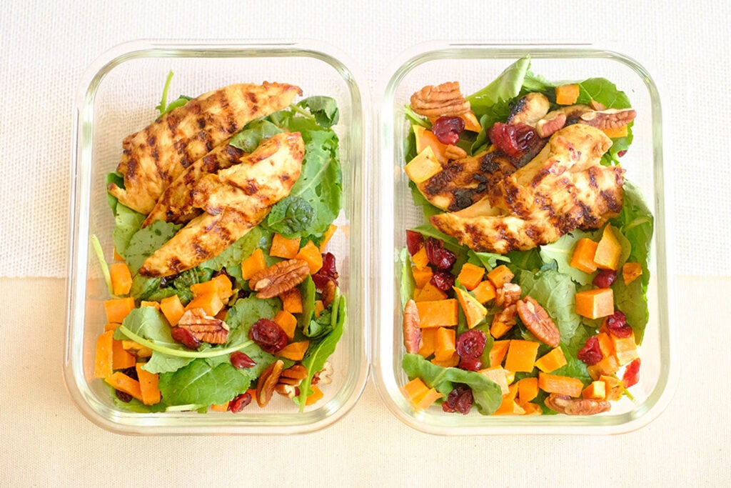 Glass meal prep containers with chicken served on top of baby kale salad with sweet potato, cranberries, and pecans.
