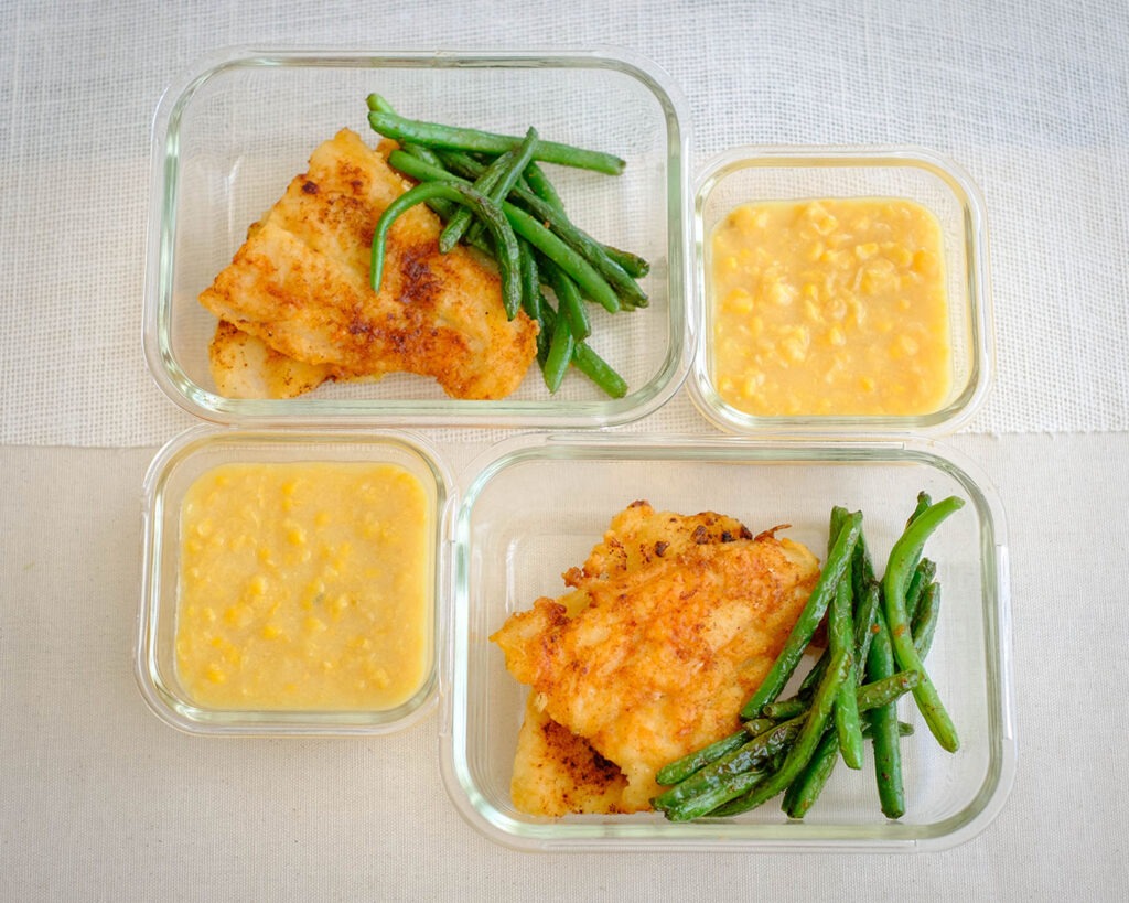 Glass meal prep containers with pan-fried fish, green beans, and creamed corn.
