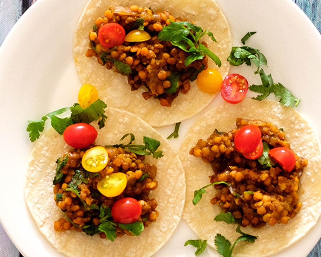 Round plate with three lentil street tacos topped with cherry tomatoes and fresh cilantro.
