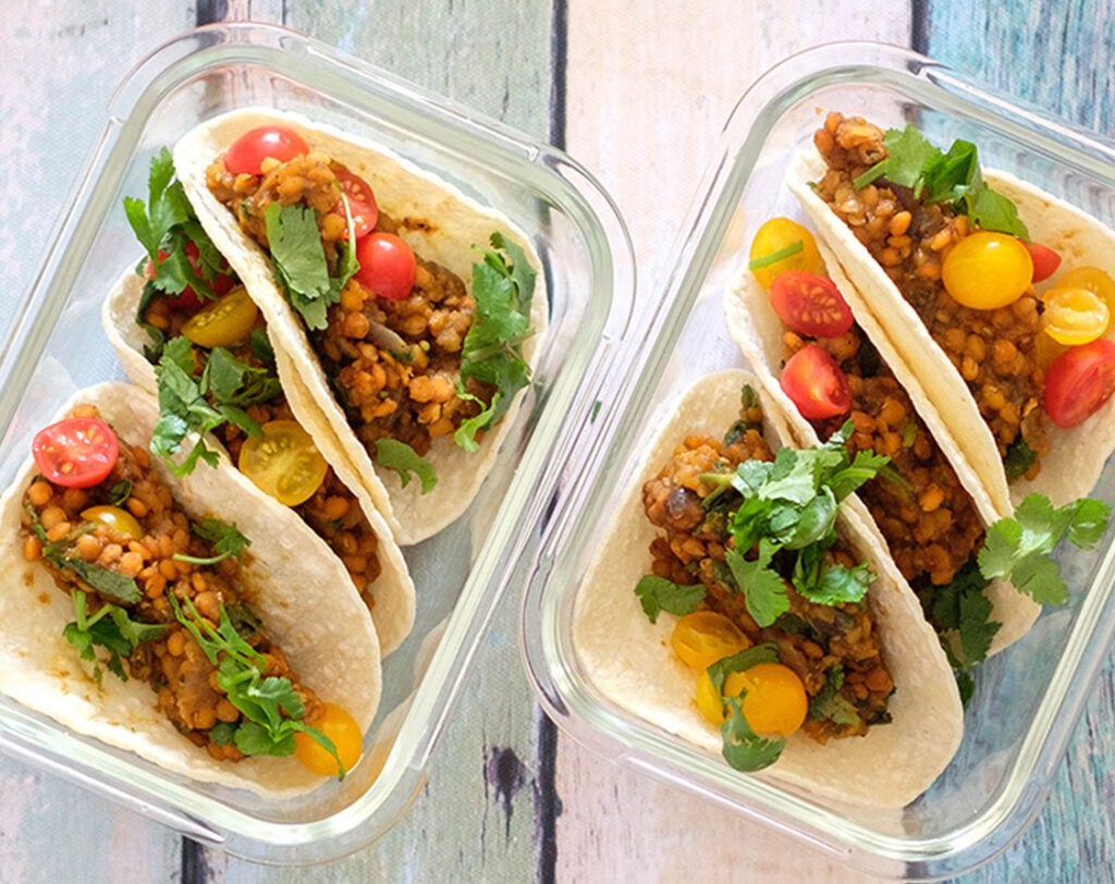 Glass meal prep containers with three lentil street tacos topped with cherry tomatoes and fresh cilantro.