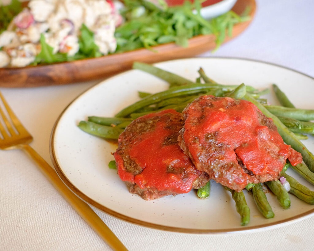 Round plate with beef patties covered in tomato sauce on top of roasted green beans.