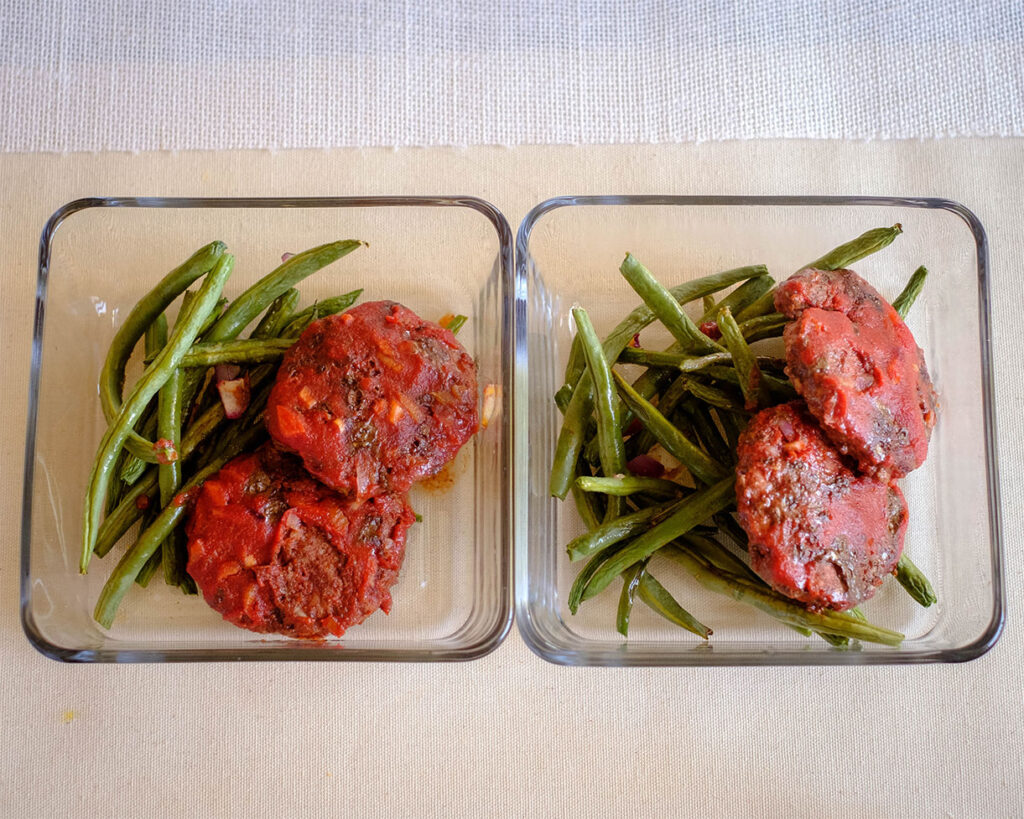 Glass meal prep containers with beef patties covered in tomato sauce on top of roasted green beans.