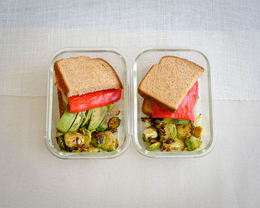 Glass meal prep containers with a grilled cheese and vegetable sandwich with roasted brussels sprouts.