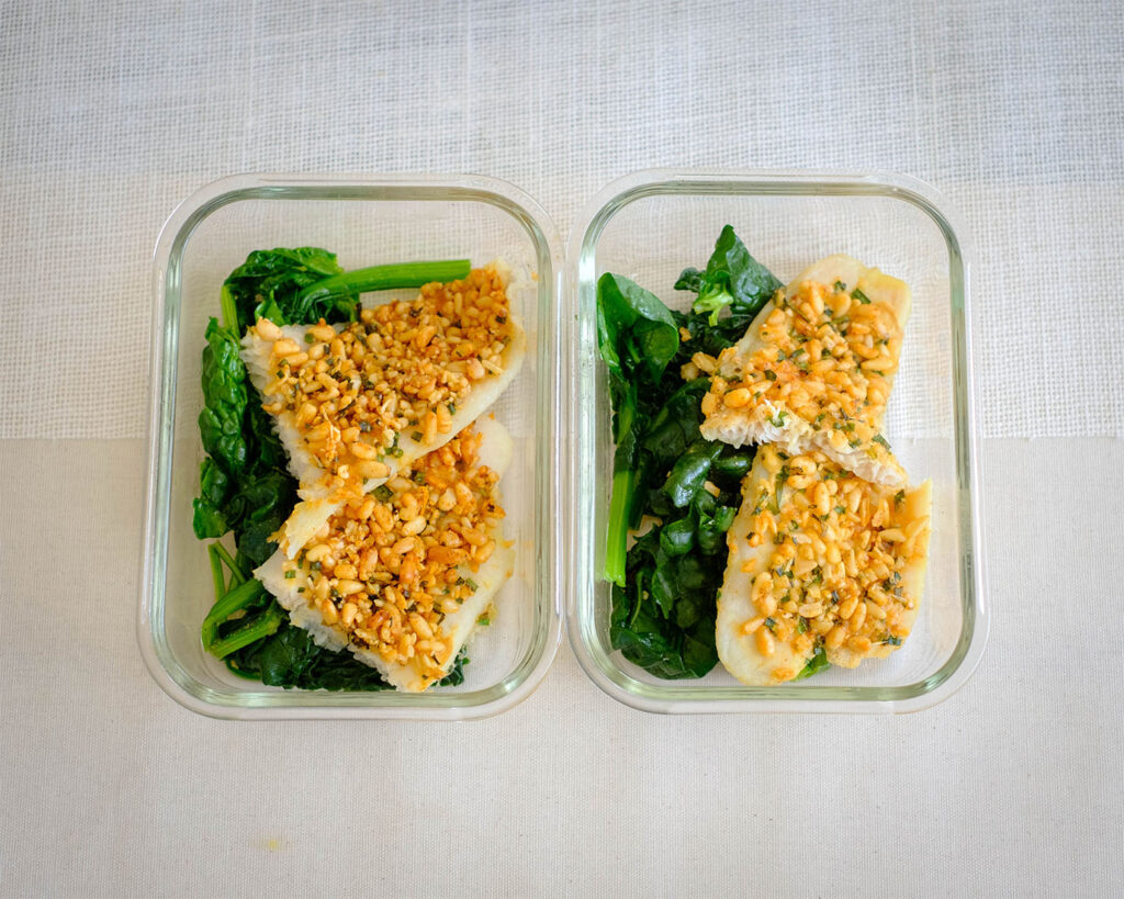 Glass meal prep containers with baked pine nut crusted tilapia with sauteed spinach.