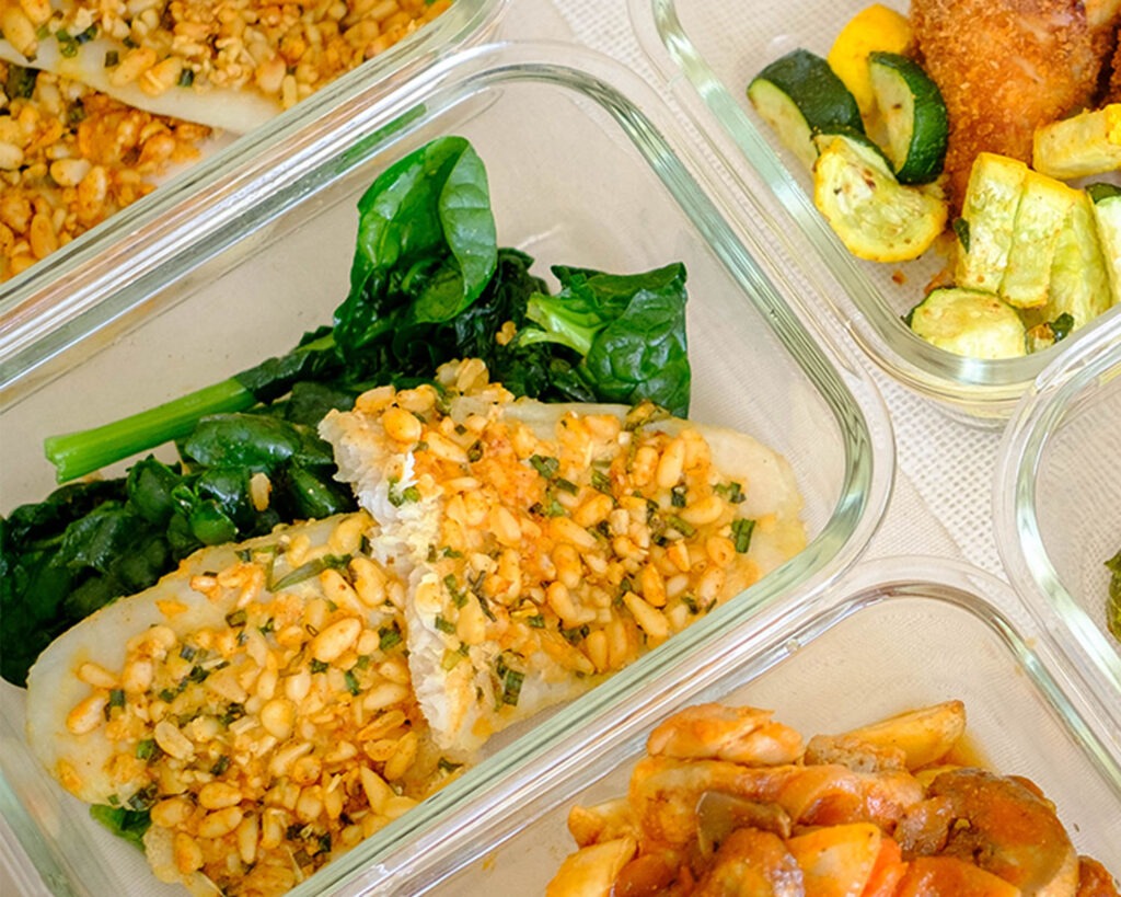 Glass meal prep container with baked pine nut crusted tilapia with sauteed spinach.