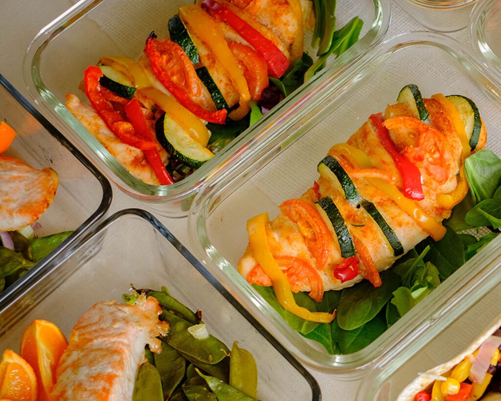 Glass meal prep containers with baked chicken breasts stuffed with colorful vegetables and a side salad.
