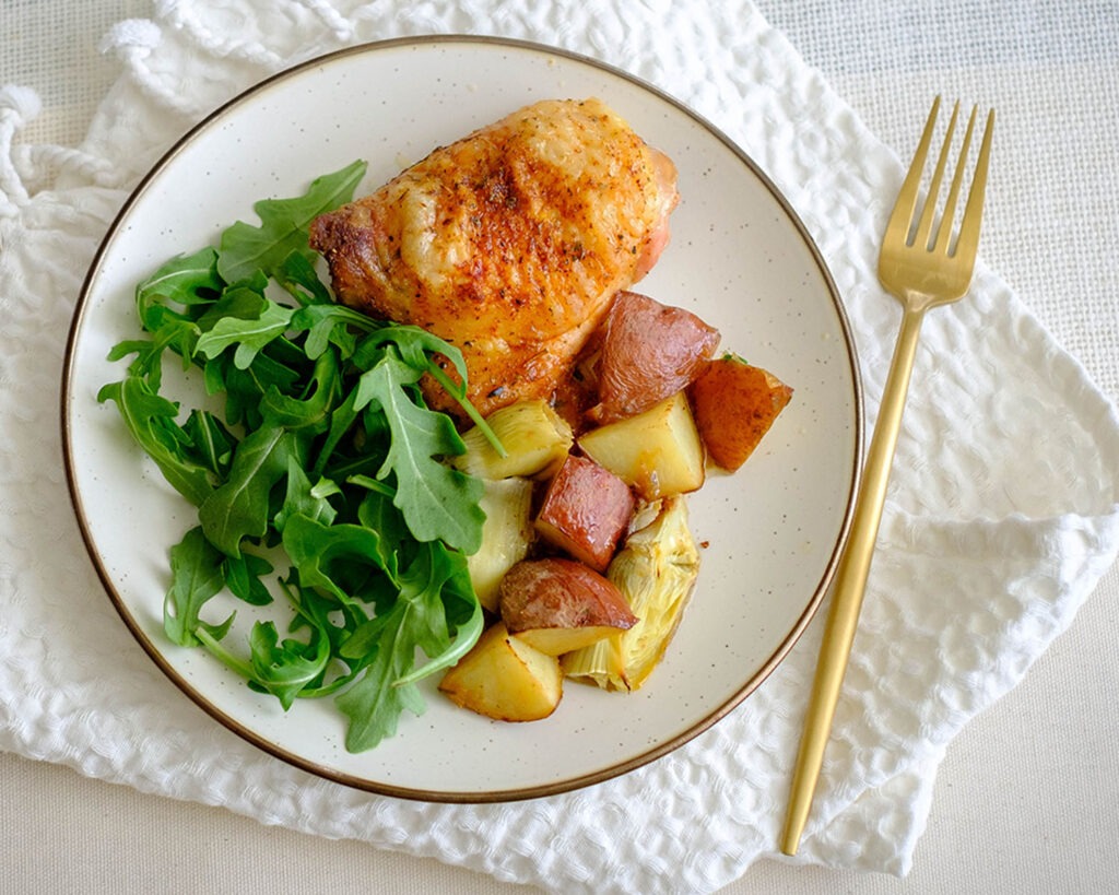 Round plate with crispy chicken thighs, roasted potatoes and artichokes served with green arugula salad.