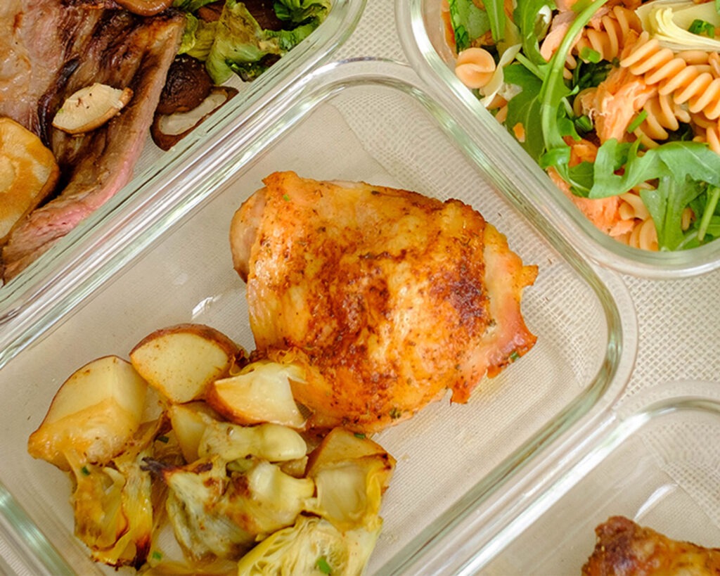 Glass meal prep container with crispy chicken thighs, roasted potatoes and artichokes.