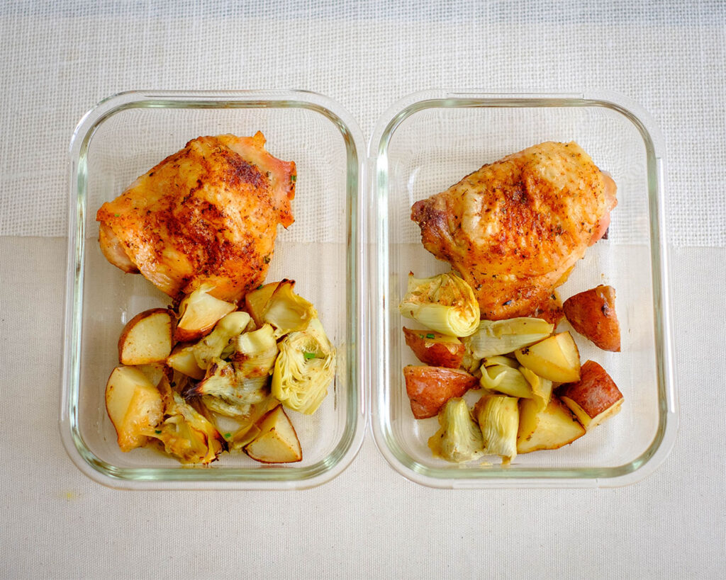Glass meal prep container with crispy chicken thighs, roasted potatoes and artichokes.