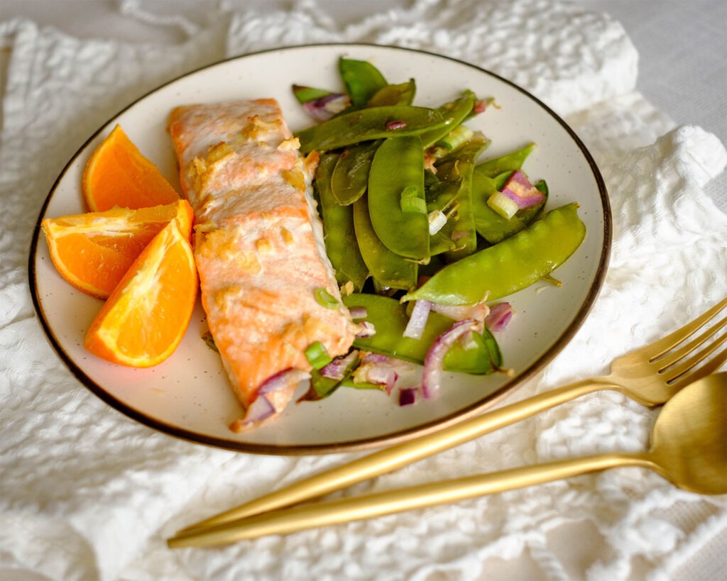Round plate with baked salmon and snow peas served with a side of sliced oranges.