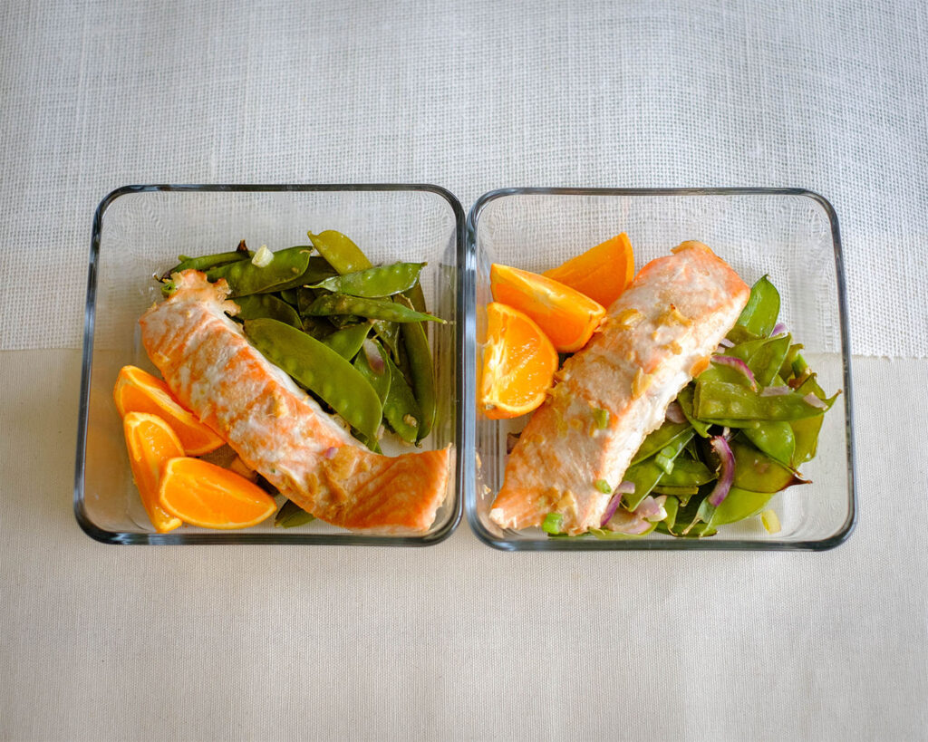 Glass meal prep containers with baked salmon and snow peas served with a side of sliced oranges.