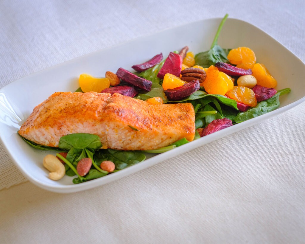 Plate with baked salmon on top of baby spinach salad with roasted beets, mandarin oranges, and nuts.