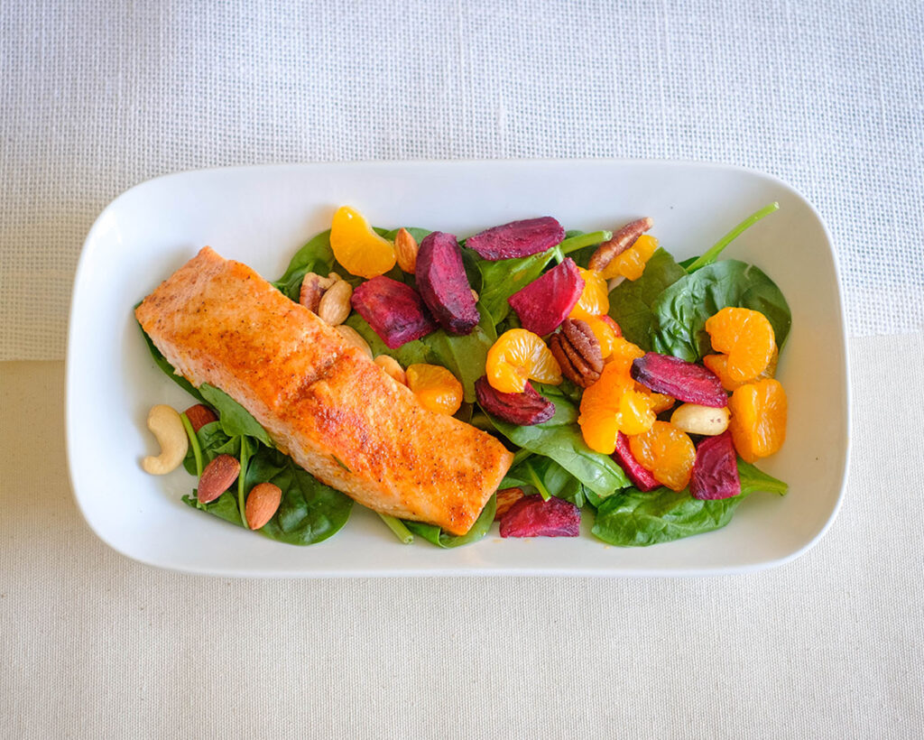 Plate with baked salmon on top of baby spinach salad with roasted beets, mandarin oranges, and nuts.