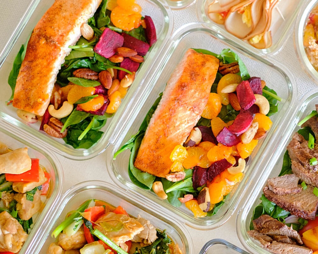 Glass meal prep containers with baked salmon on top of baby spinach salad with roasted beets, mandarin oranges, and nuts.