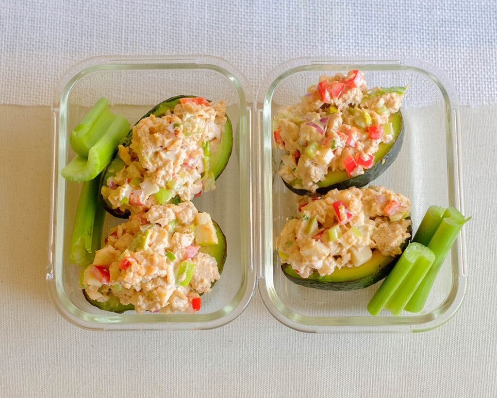 Glass meal prep containers with avocado stuffed with salmon salad and celery sticks.