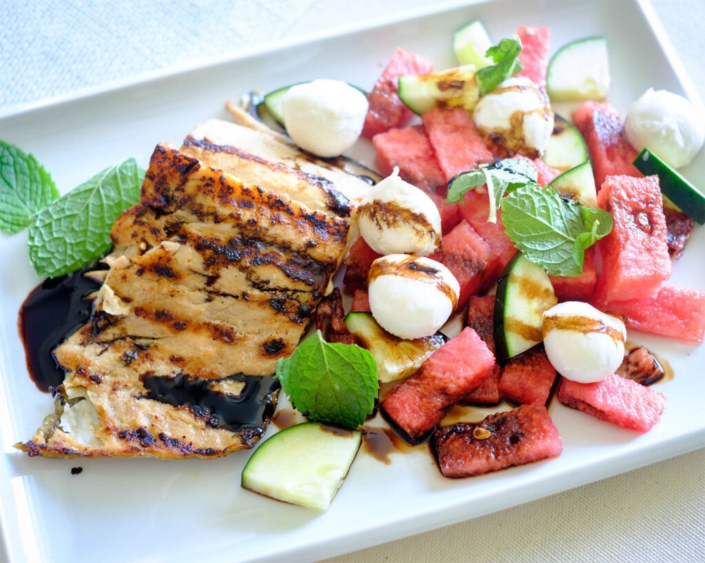 Plate with grilled salmon and watermelon salad with mozzarella balls, cucumbers, and mint leaves drizzled with balsamic glaze.