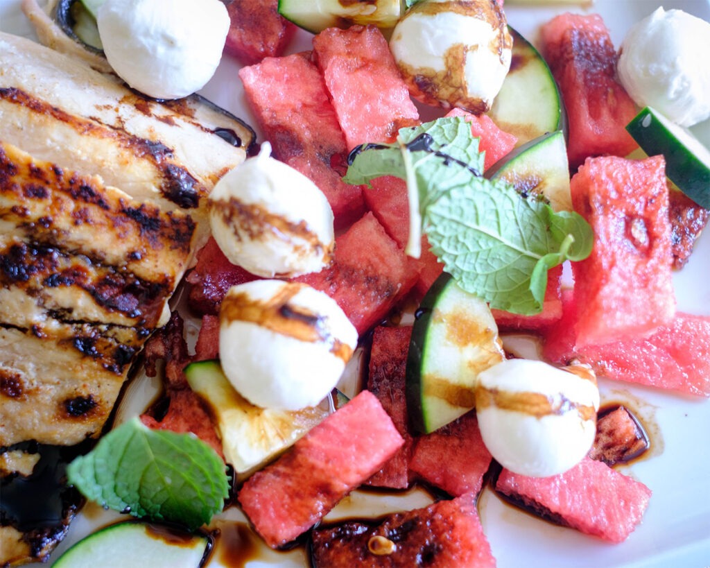 Plate with grilled salmon and watermelon salad with mozzarella balls, cucumbers, and mint leaves drizzled with balsamic glaze.