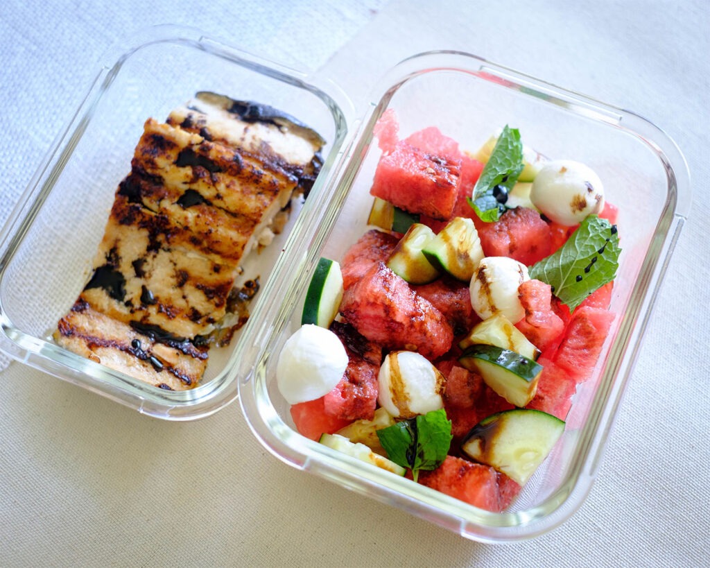 Glass meal prep containers with grilled salmon and watermelon salad with mozzarella balls, cucumbers, and mint leaves drizzled with balsamic glaze.