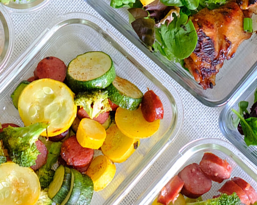 Glass meal prep containers with roasted sausage, zucchini, yellow squash, and broccoli florets.