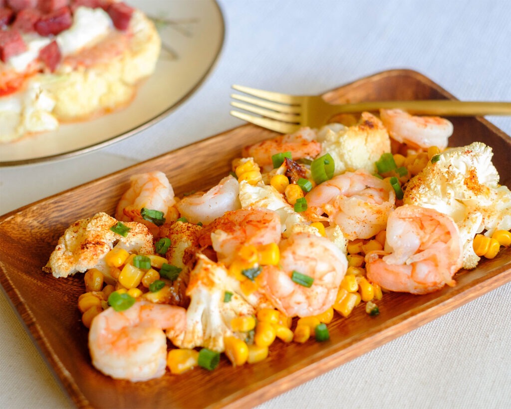 Wood plate with roasted shrimp, corn, cauliflower, and green onions.