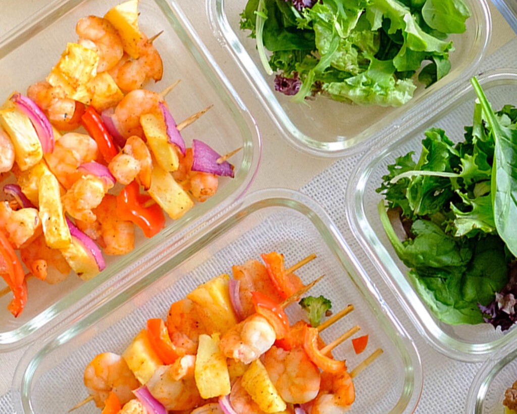 Glass meal prep containers with colorful shrimp kebabs that include shrimp, pineapple, red onions, and red bell pepper.