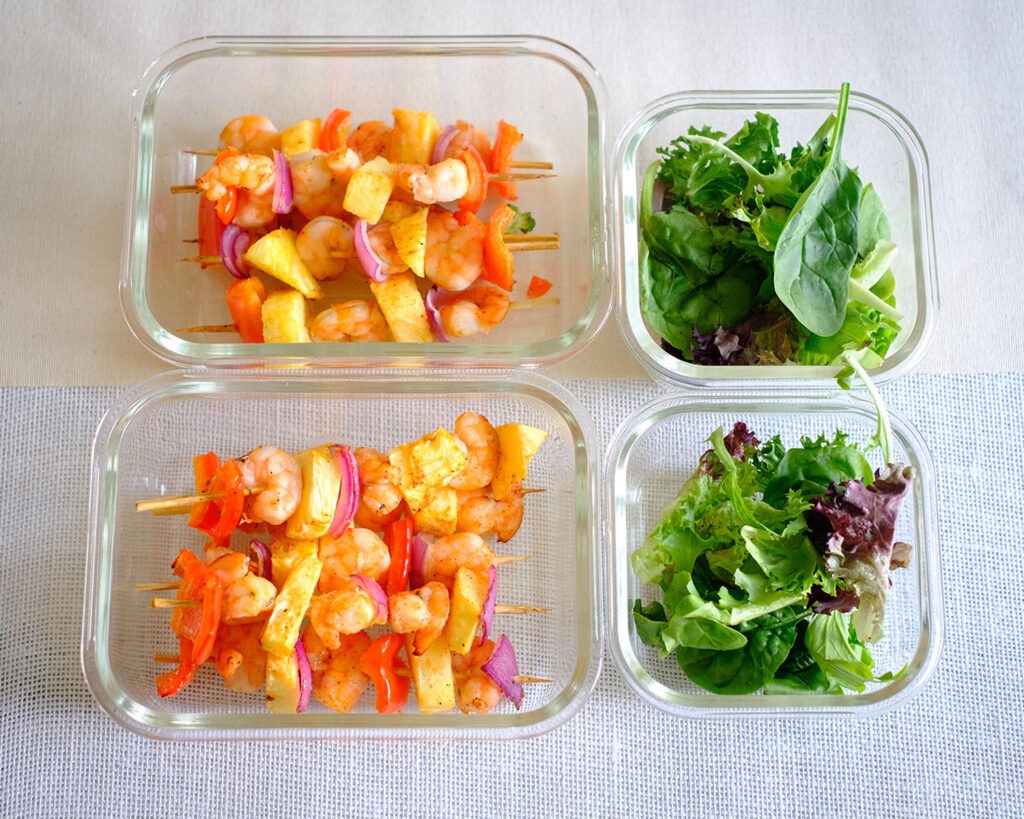 Glass meal prep containers with colorful shrimp kebabs that include shrimp, pineapple, red onions, and red bell pepper. Included is a side salad.