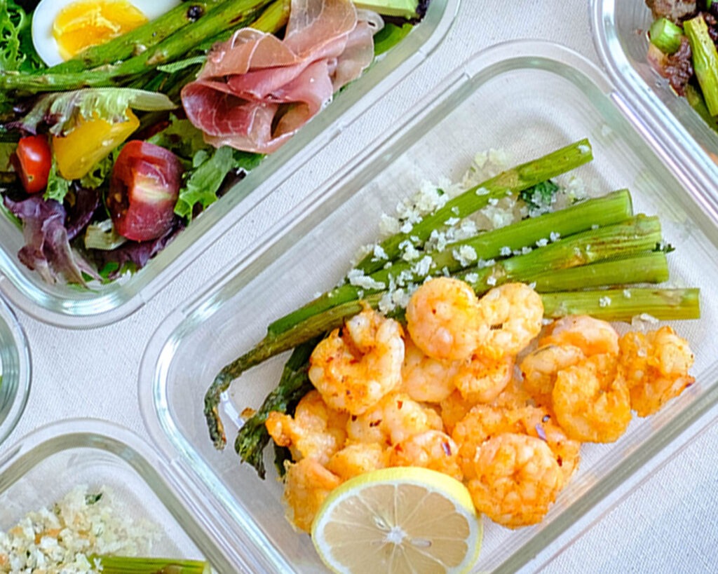 Glass meal prep container with roasted shrimp and asparagus topped with panko breadcrumbs and lemon slice.