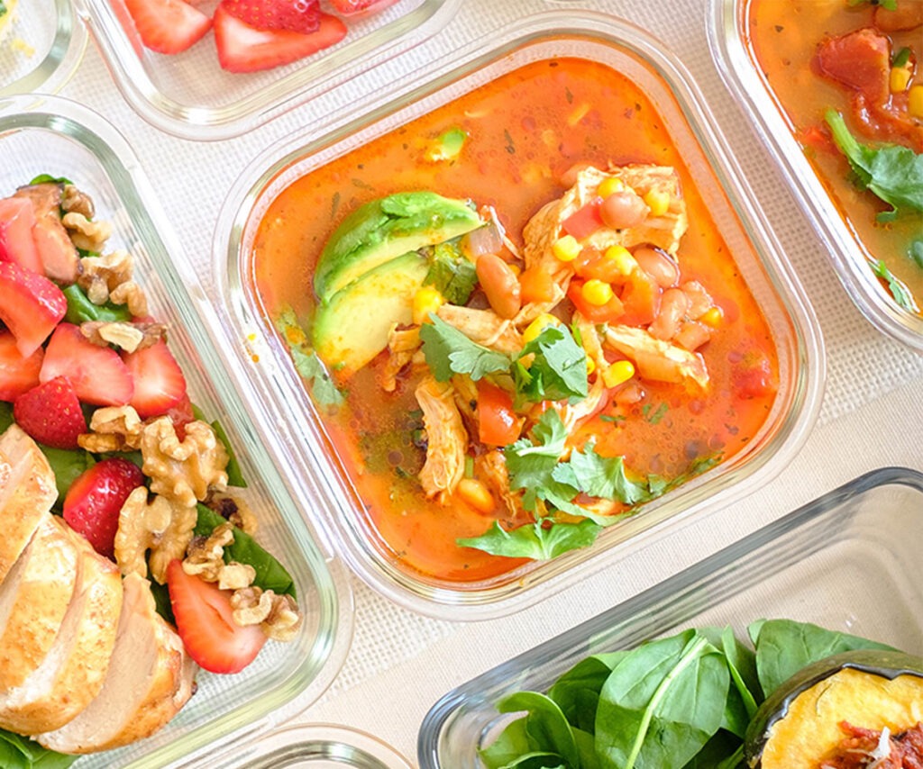 Glass meal prep container of chicken soup with beans, vegetables, avocado, and cilantro