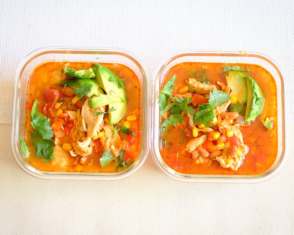 Glass meal prep containers of chicken soup with beans, vegetables, avocado, and cilantro