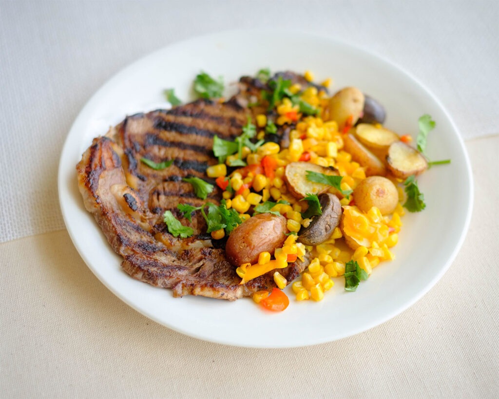 Round plate with grilled ribeye steak with corn, baby potatoes, and bell peppers.