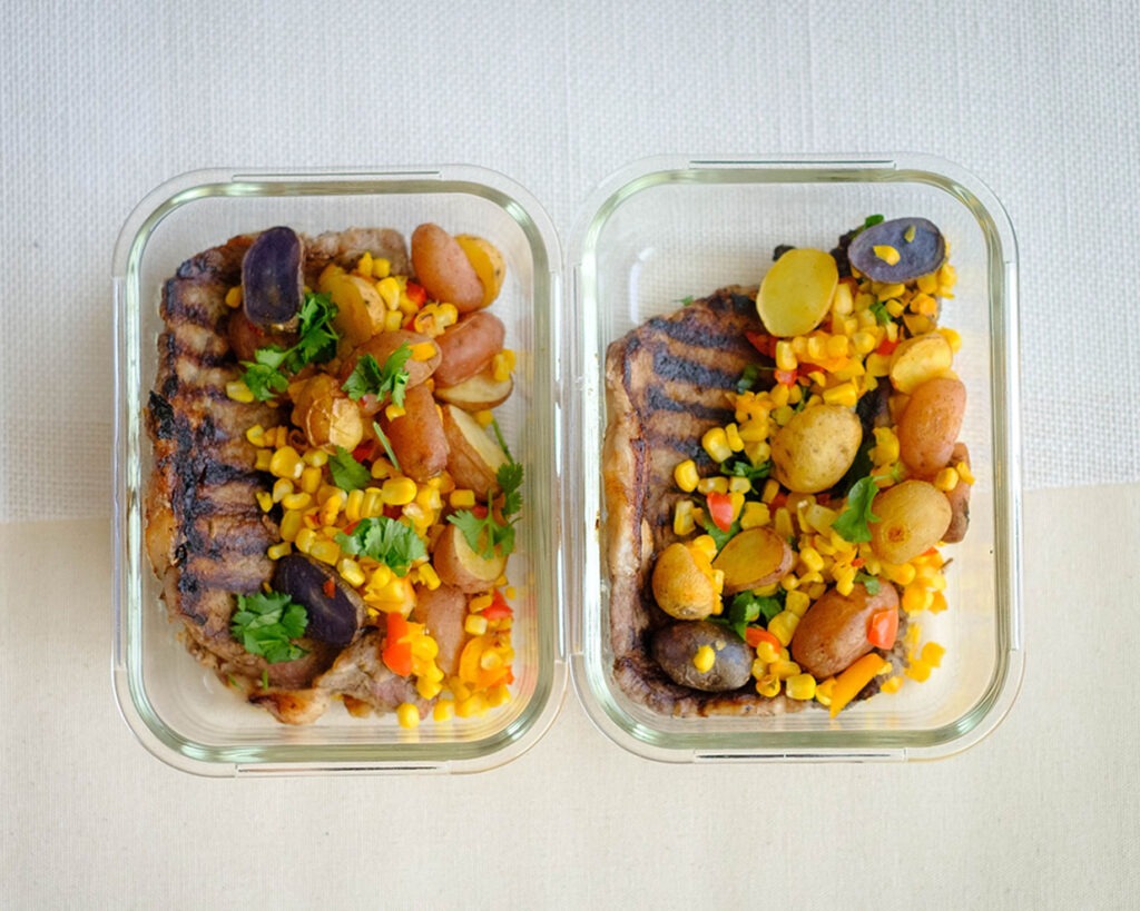 Glass meal prep container with grilled ribeye steak with corn, baby potatoes, and bell peppers.