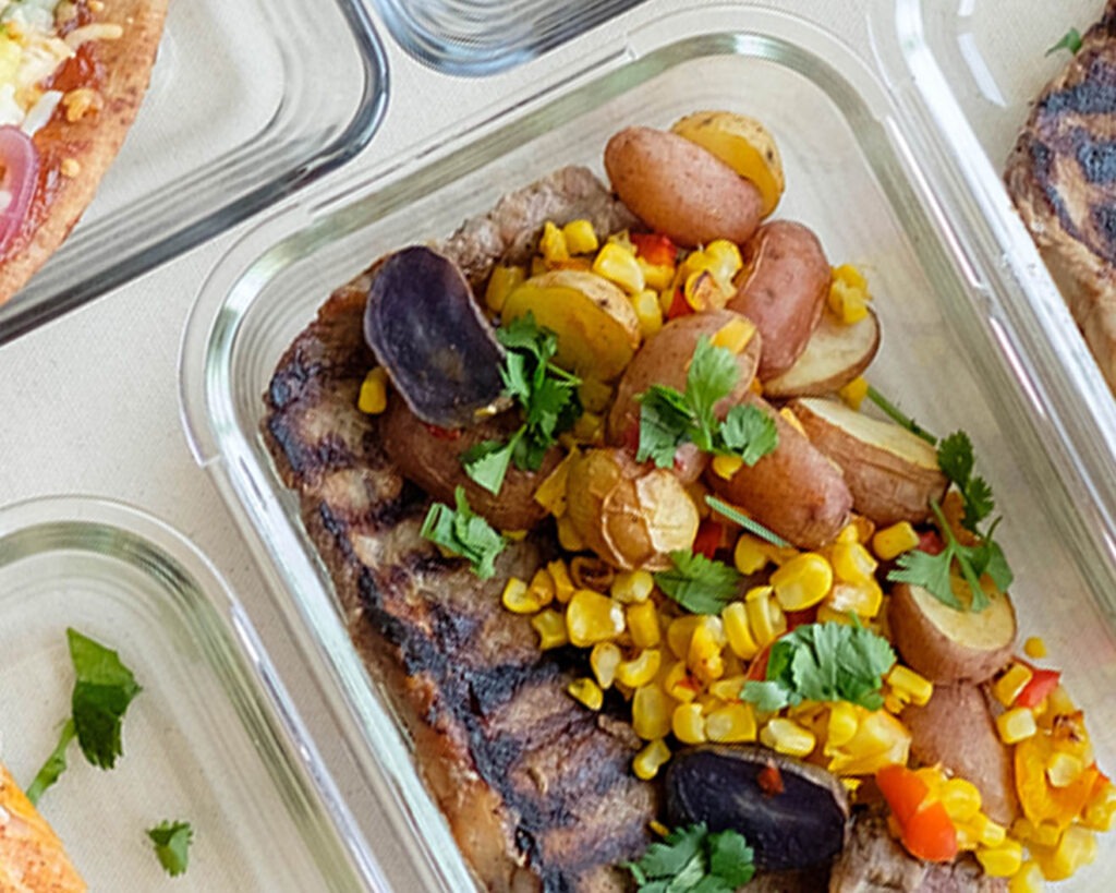 Glass meal prep container with grilled ribeye steak with corn, baby potatoes, and bell peppers.